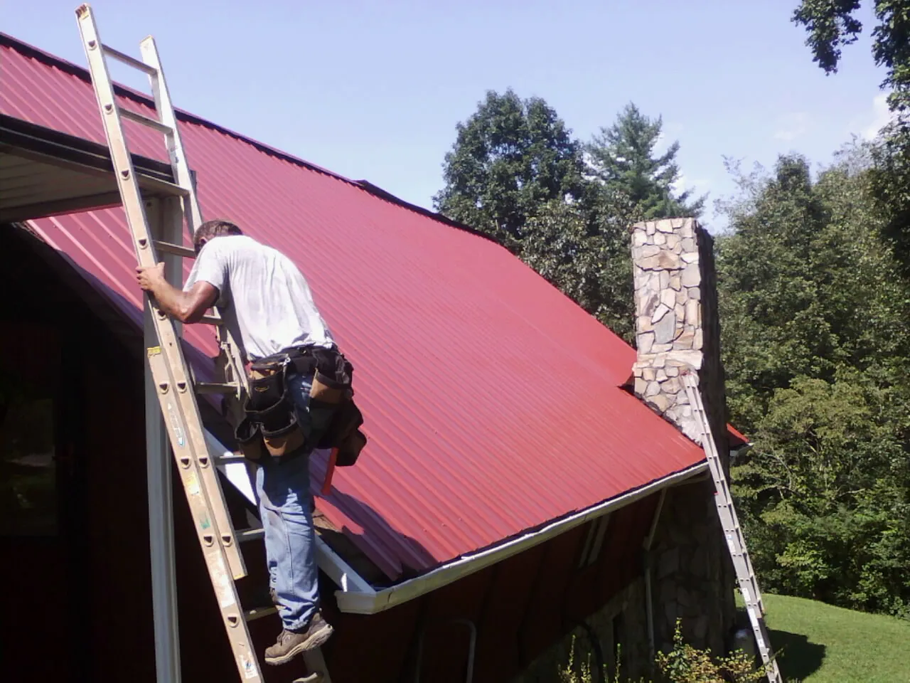 Roofer on a ladder beside a red metal roof and stone chimney — Soffit & Fascia Repair by Roofing Brothers in Gerrardstown